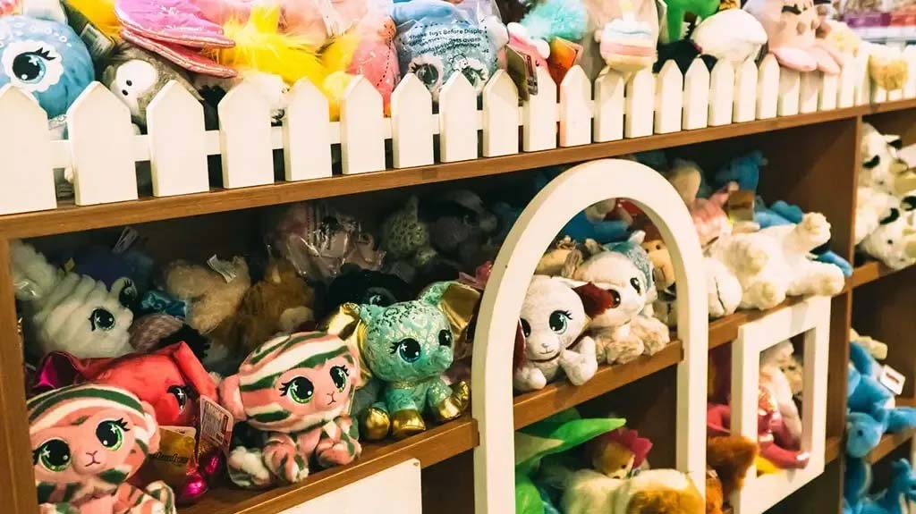 A colorful variety of plush toys neatly arranged on wooden display shelves with a white picket fence design, featuring stuffed animals and cute character plushies in a toy store.
