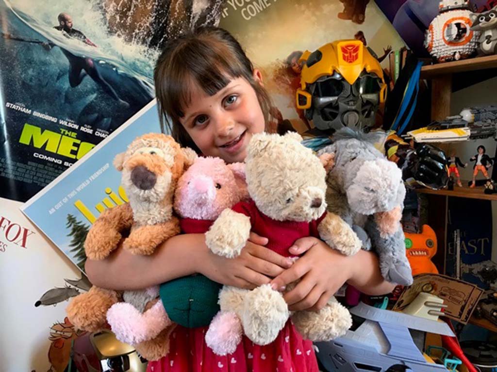 Smiling young girl in a red dress holding several Winnie the Pooh plush toys, including Pooh, Piglet, Tigger, and Eeyore, in a cozy room decorated with movie posters and toys.