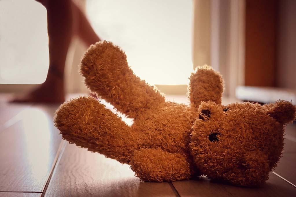 Brown teddy bear plush toy lying on a wooden floor in warm sunlight, with a person walking in the background near a doorway.