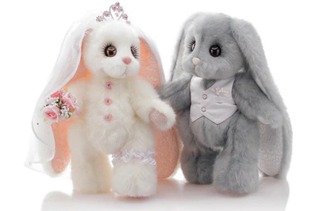 Two plush bunny toys dressed as a bride and groom, with the white bunny wearing a veil and holding a bouquet, and the gray bunny in a vest suit.