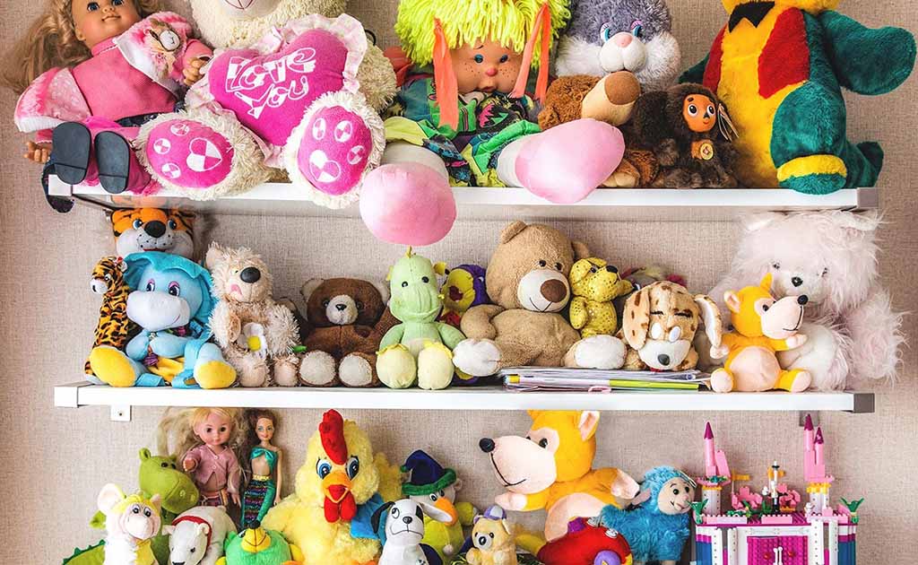 A vibrant assortment of plush toys, teddy bears, dolls, and stuffed animals arranged neatly on white shelves against a beige wall.