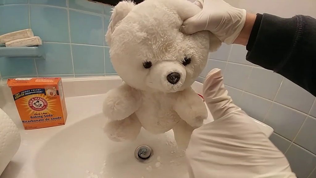 Person wearing gloves cleaning a white teddy bear in a sink using baking soda, with a box of Arm & Hammer baking soda visible in the background.