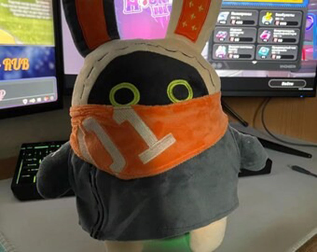 Cute black bunny plush with orange ears and a matching orange scarf printed with the number 01, dressed in a gray hoodie and placed on a desk near computer monitors.