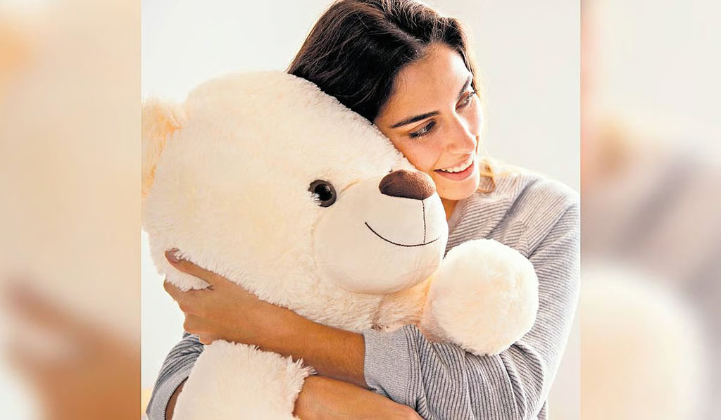 Smiling woman in a light sweater warmly hugging a big cream-colored teddy bear in a bright, cozy room.