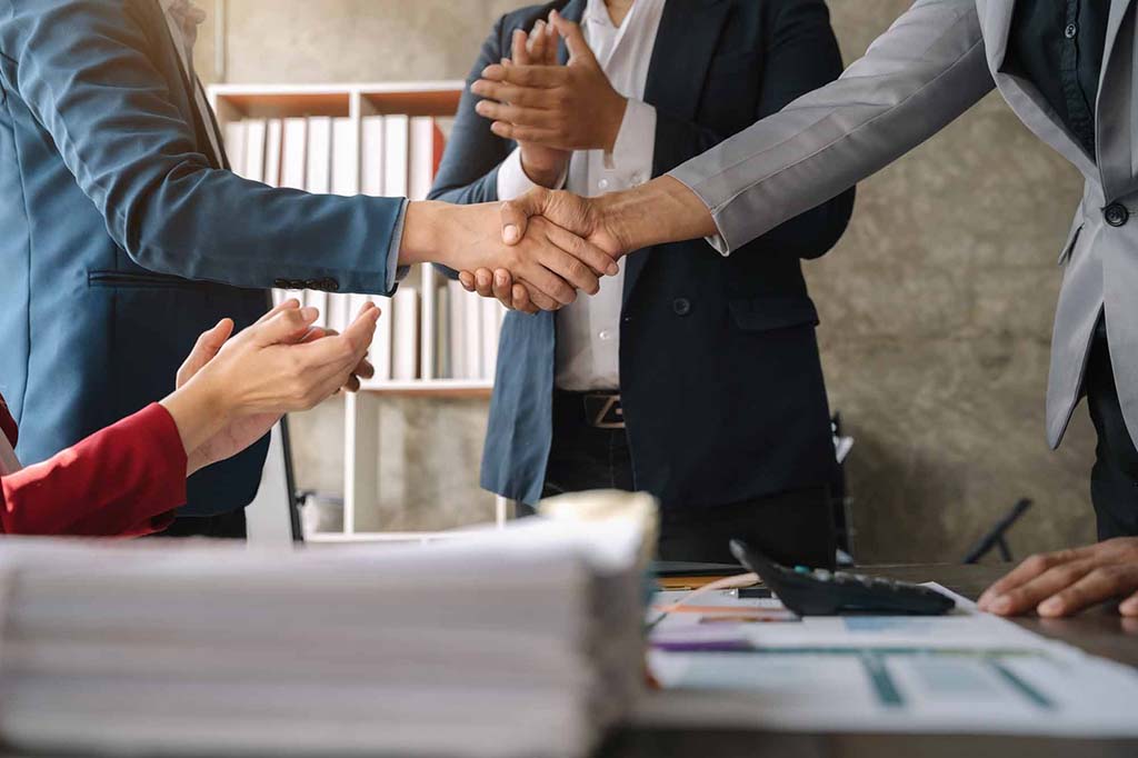 Business professionals in formal suits shaking hands and clapping around a meeting table, symbolizing successful partnership or agreement.