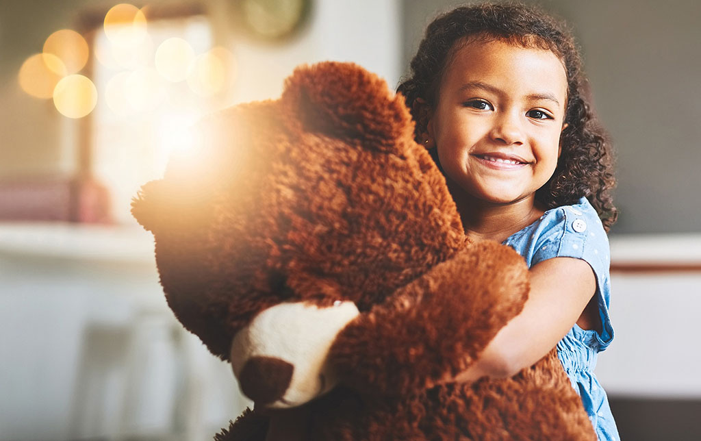Smiling young girl in a blue dress hugging a large brown teddy bear with warm sunlight shining in the background.