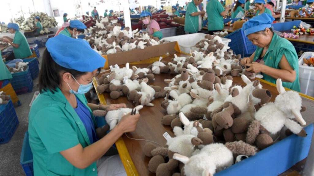 Factory workers wearing blue caps assembling and sewing brown and white plush toys on a production line.