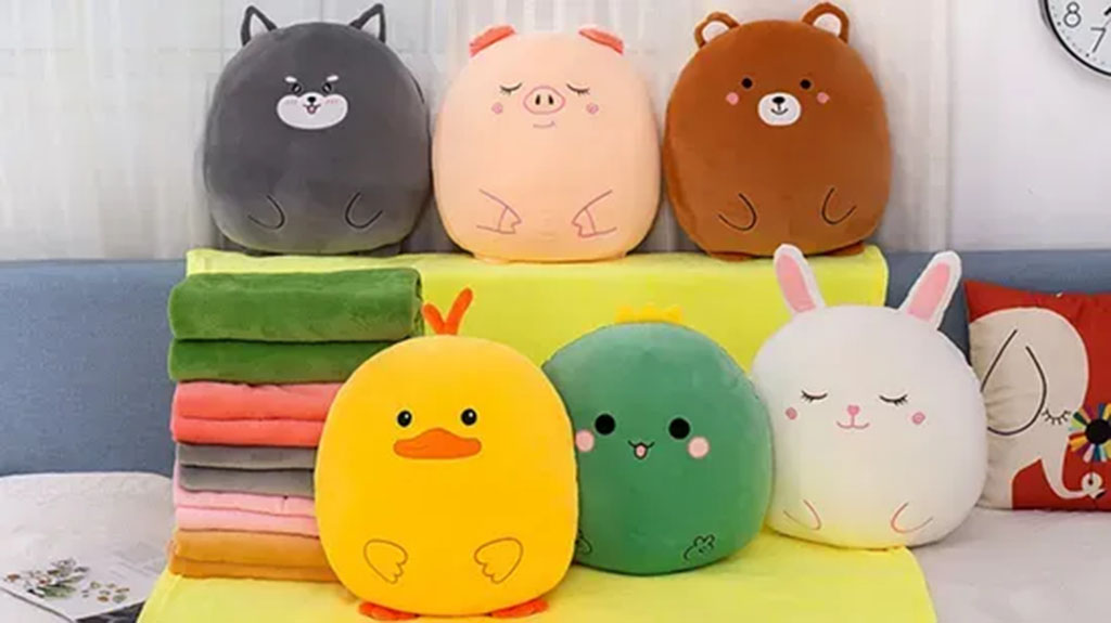 Soft round plush pillows shaped like animals—including a duck, rabbit, bear, pig, cat, and green pea—displayed with folded colorful blankets on a couch.