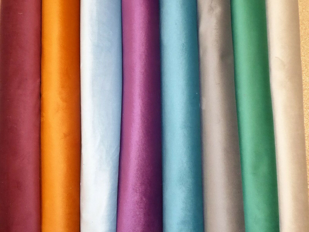 Multiple rolls of soft plush velvet fabric in various rich colors, including gold, purple, teal, and green.