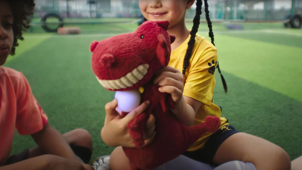 Smiling child holding a red dinosaur plush toy on a green playground, with another child sitting nearby.