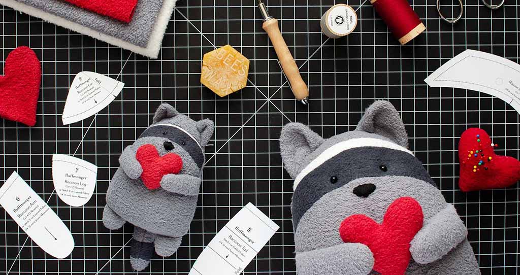 Two gray raccoon plush toys holding red hearts on a crafting grid surrounded by sewing tools, fabric pieces, and pattern templates.