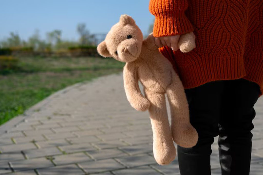 Child wearing an orange sweater and black pants holding a soft brown teddy bear by one arm while walking on a paved path in a park.