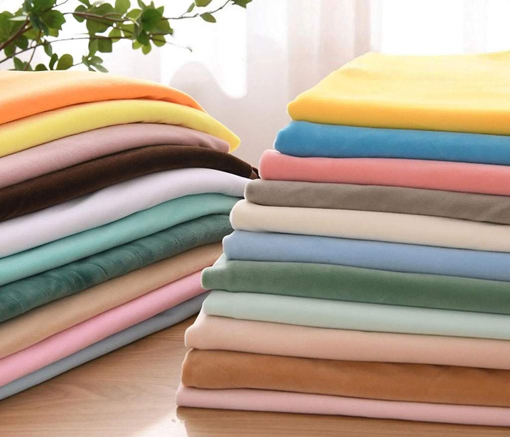 Neatly folded stacks of soft plush fabrics in pastel and vibrant colors on a wooden table near sunlight.