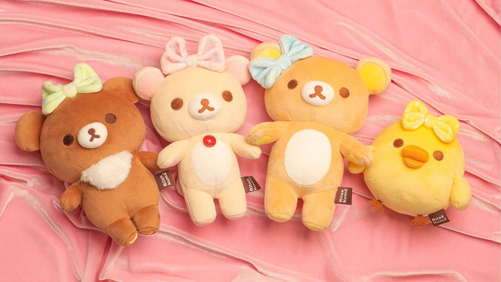 Four cute Rilakkuma plush toys, including bears and a chick, each wearing pastel bows, displayed on a soft pink fabric background.