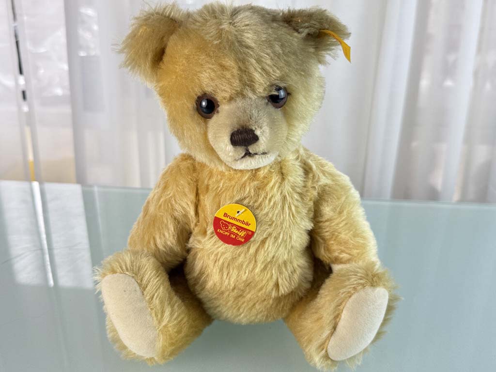 A classic light brown Steiff teddy bear with a yellow ear tag and red chest label, sitting on a glass surface in front of sheer curtains.