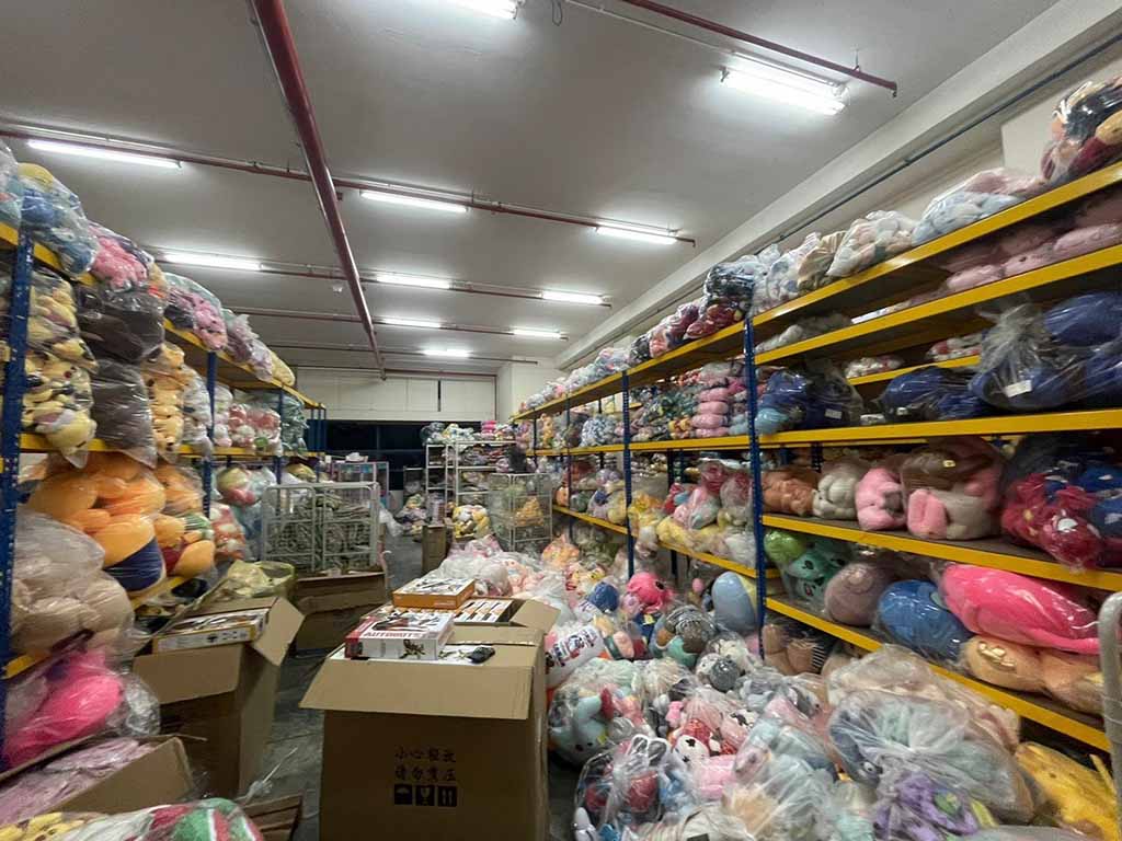Shelves filled with bagged plush toys in a brightly lit warehouse, with cardboard boxes and packaged stuffed animals ready for distribution.