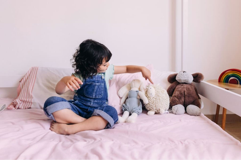 Young child in denim overalls sitting on a pink bed, arranging and playing with three plush toys — a bunny, a sheep, and a bear — in a cozy bedroom setting.