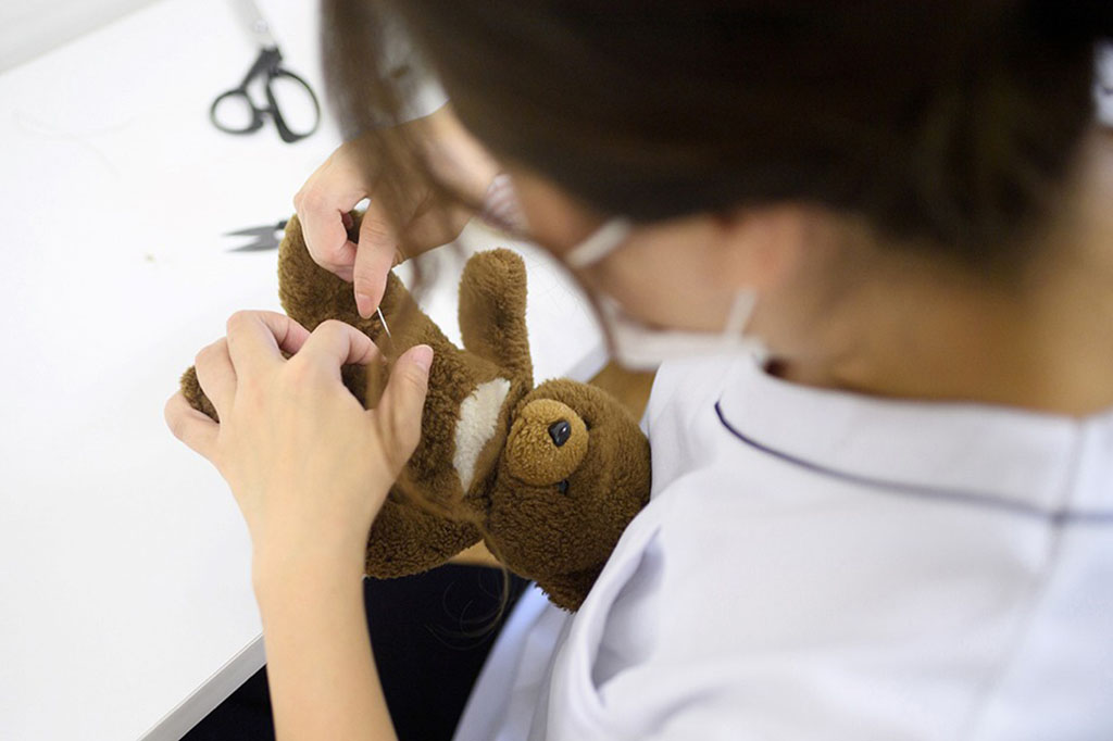 Worker in white uniform hand-sewing a brown teddy bear on a table with scissors and thread nearby.