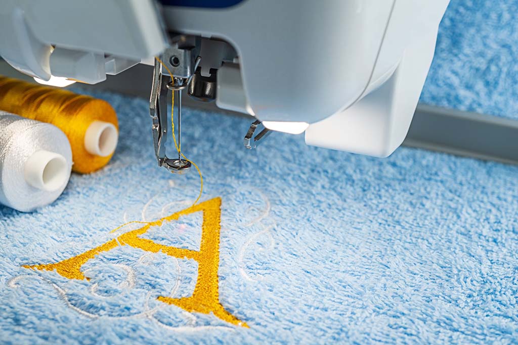 Close-up of embroidery machine sewing yellow letter A on blue fabric with thread spools nearby.