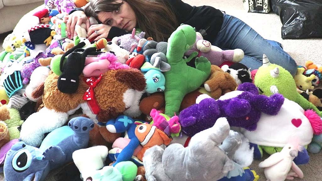 Woman lying on a carpet surrounded by a large pile of colorful plush toys, including animals and cartoon characters, showing a mix of soft textures and vibrant colors.