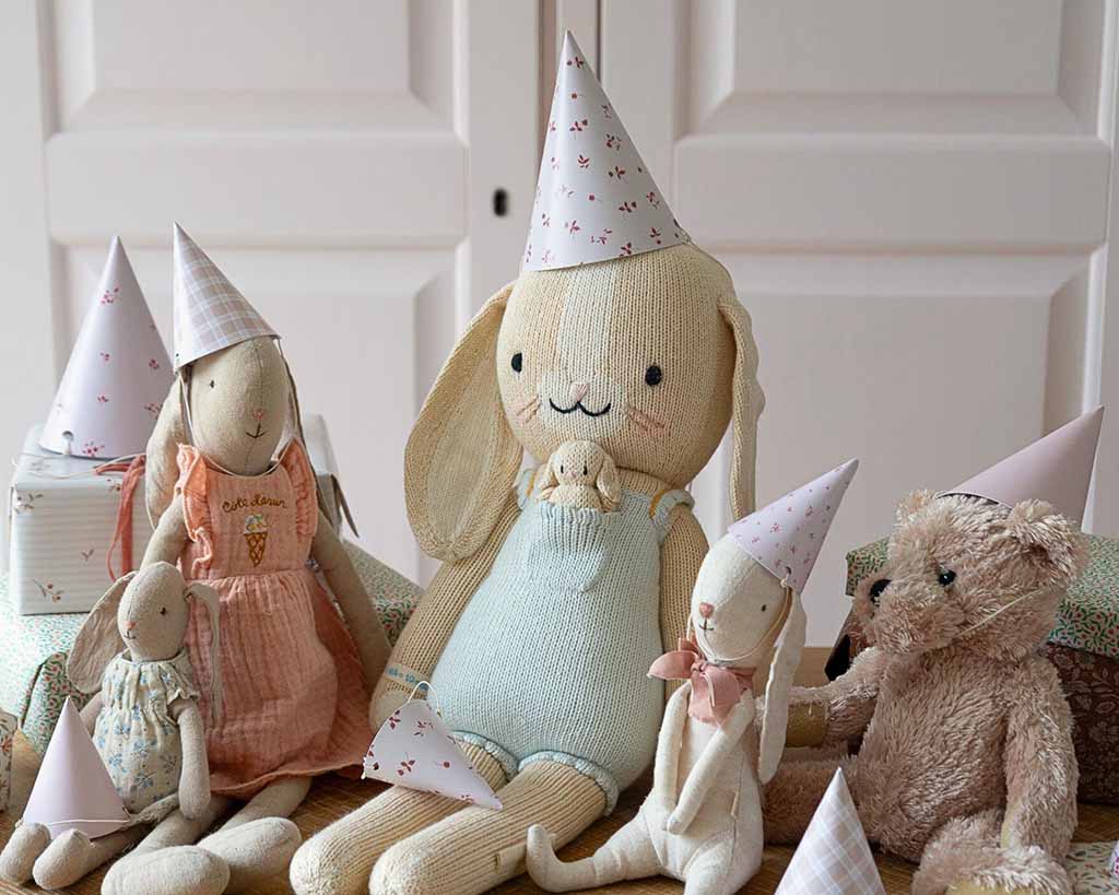 A group of soft plush animals, including rabbits and a teddy bear, wearing pastel party hats and sitting together as if celebrating a toy tea party.