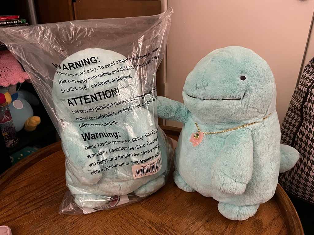 A soft mint-green plush toy sitting beside another identical one still sealed in a plastic warning bag on a wooden table.