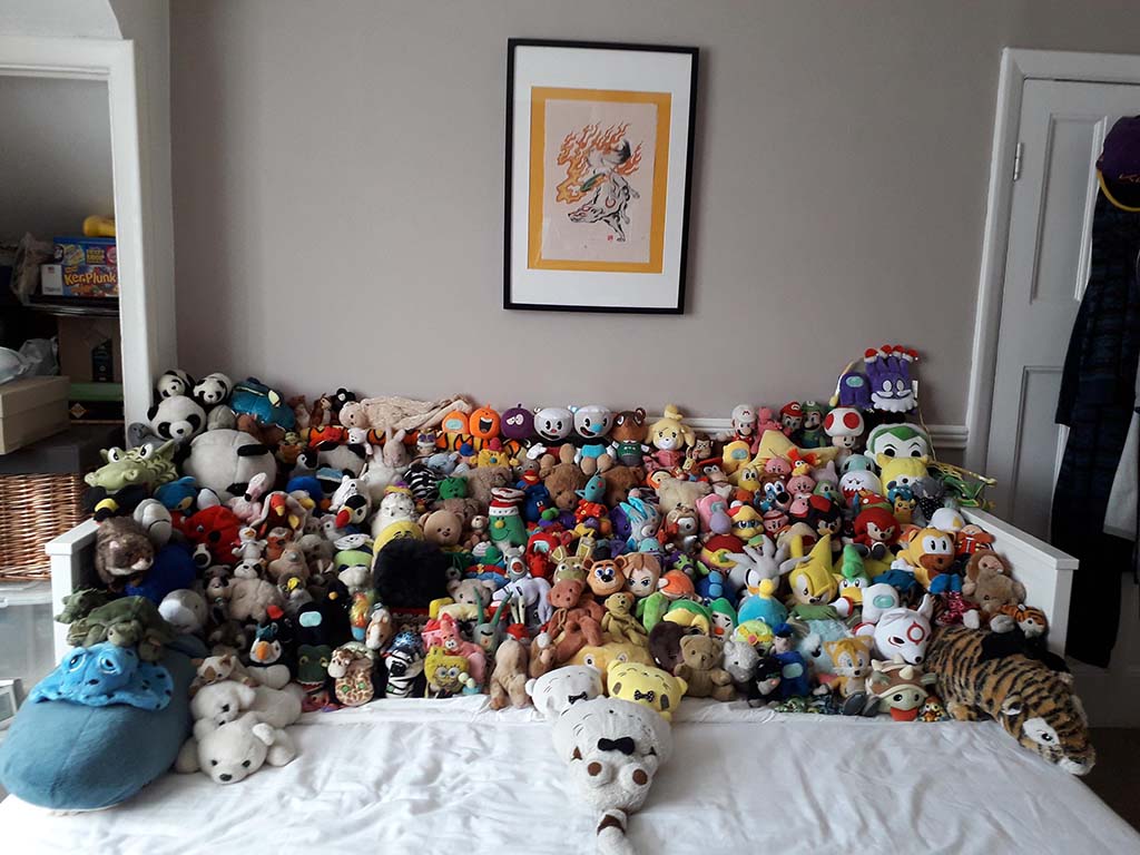 A large collection of colorful plush toys and stuffed animals arranged across a bed beneath a framed artwork on a beige wall.