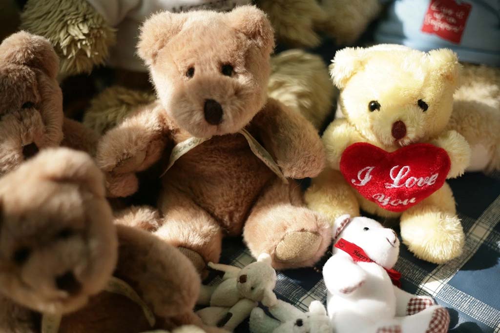 A group of teddy bears, including a yellow one holding a red heart with “I Love You” embroidered on it, arranged on a checkered surface in warm lighting.