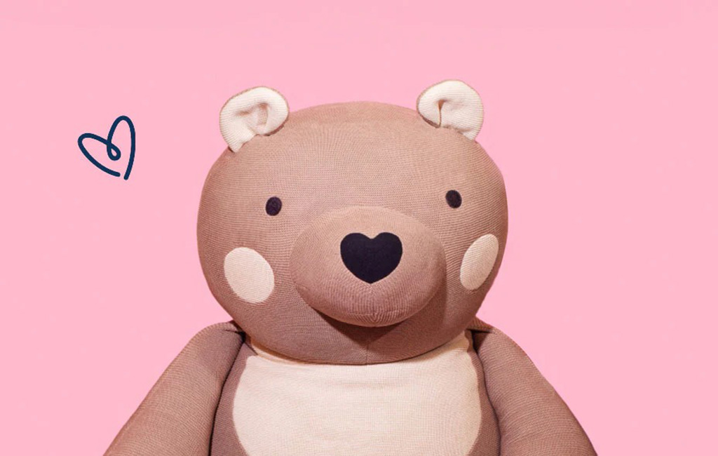 A soft brown plush bear with a heart-shaped nose and blush cheeks sitting against a pink background, symbolizing love and comfort.