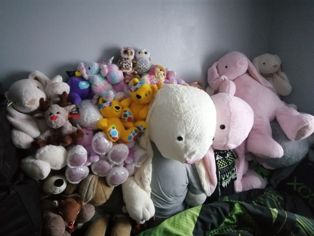 A colorful assortment of plush toys, including large bunnies, bears, unicorns, reindeer, and owls, piled together on a bed against a gray wall.