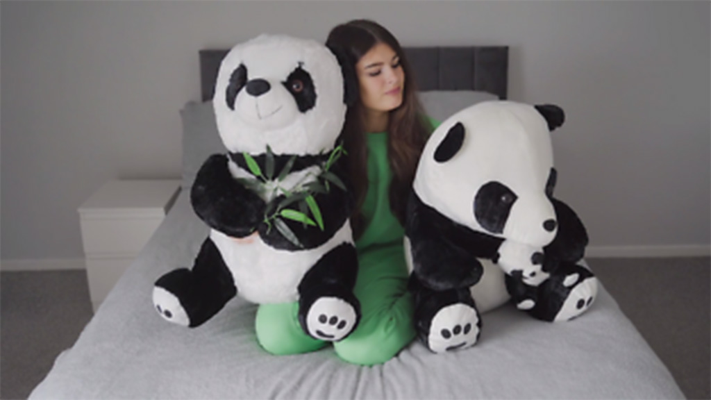 A young woman sitting on a bed between two large panda plush toys, one holding bamboo leaves, in a minimal gray bedroom setting.