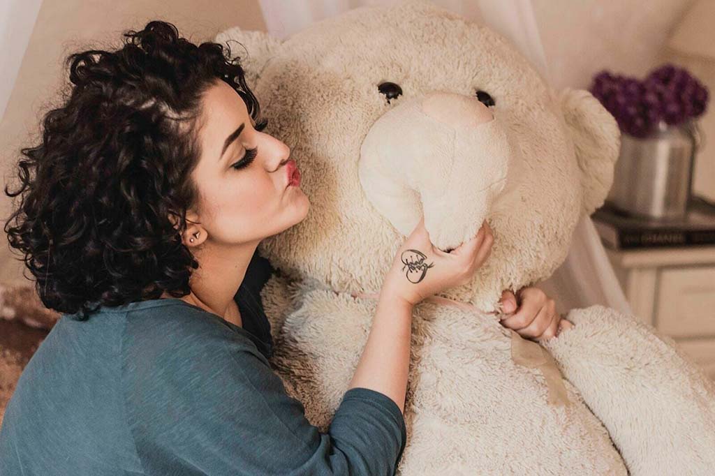 A woman with curly hair playfully kissing the cheek of a giant cream-colored teddy bear while sitting indoors.