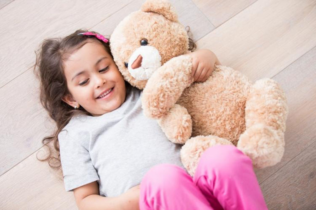 Smiling young girl lying on a wooden floor in a gray shirt and pink pants, cuddling a soft brown teddy bear.