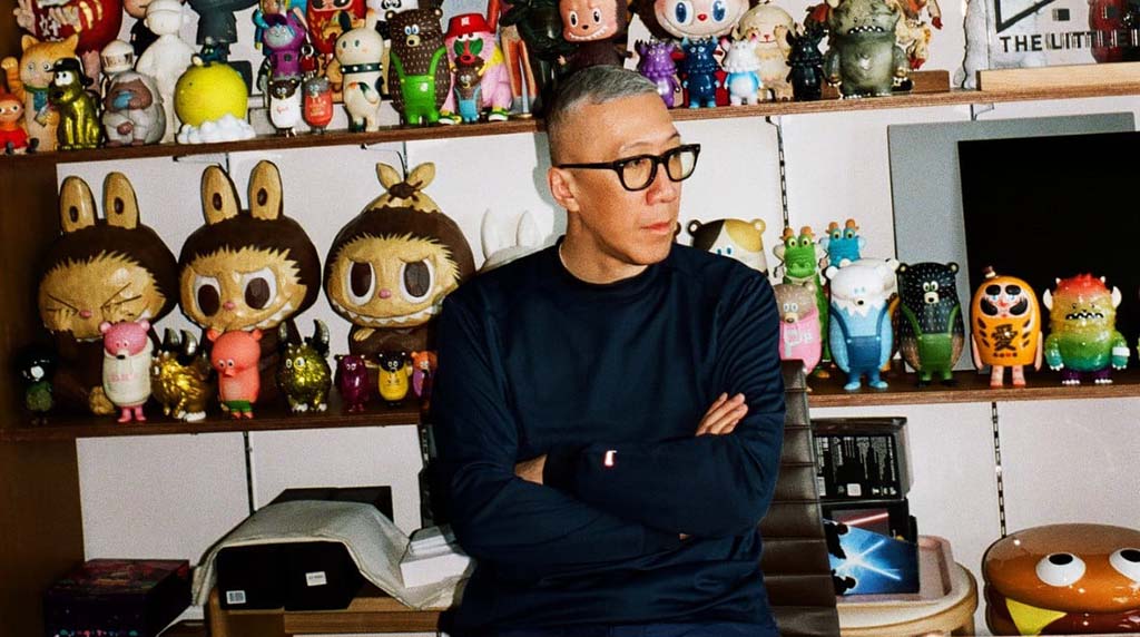 Man in glasses sitting with arms crossed in front of shelves filled with colorful designer vinyl and plush toys in a creative studio setting.