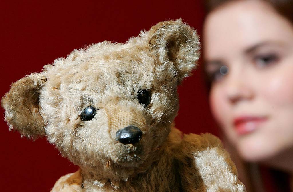 A close-up of an old, worn teddy bear with glass eyes and a stitched nose, with a woman blurred in the background against a red wall.