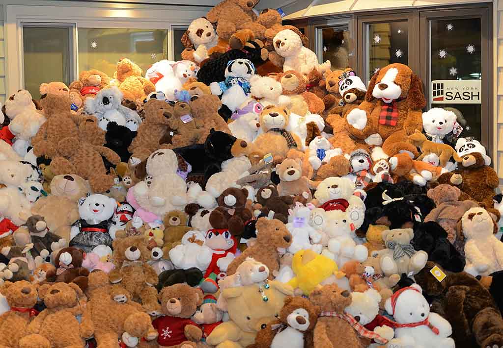 A large pile of assorted teddy bears and plush animals stacked in front of a building window, creating a mountain of stuffed toys.