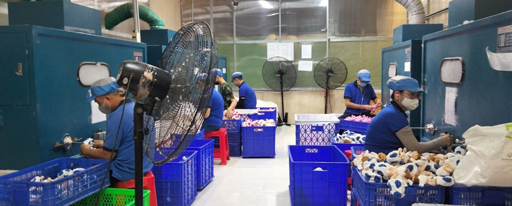 Factory workers wearing masks and blue uniforms operating plush toy drying and sterilization machines, with bins full of stuffed animals ready for inspection.
