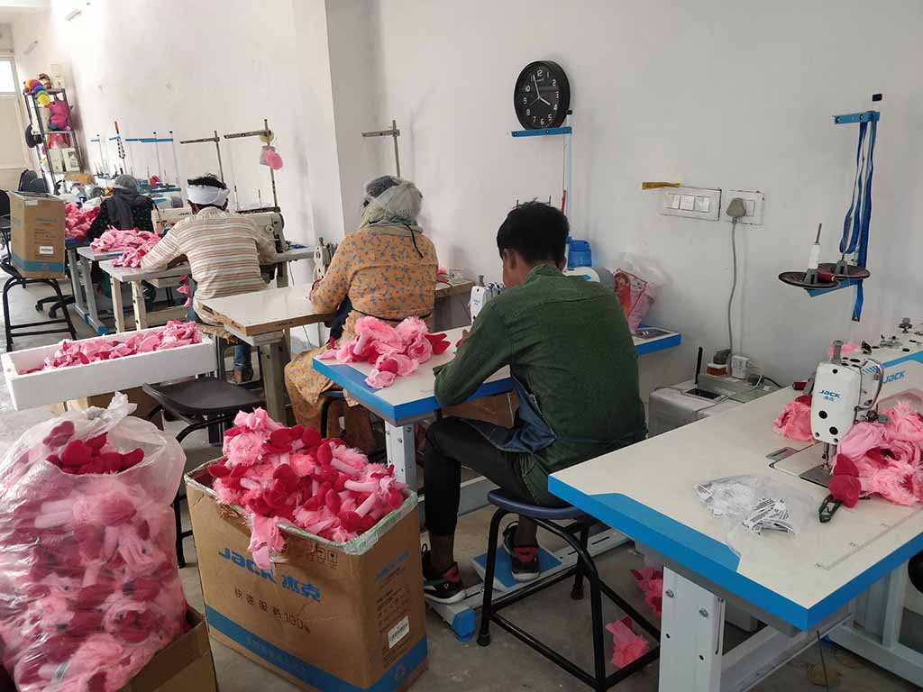 Factory workers stitching pink plush toy parts at sewing stations in a stuffed toy manufacturing workshop.