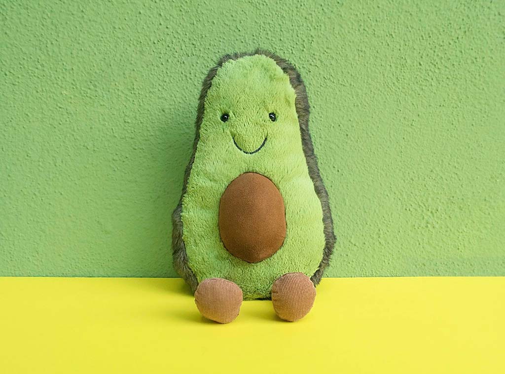 A smiling avocado plush toy with brown feet and a soft green body, sitting against a matching green and yellow background.