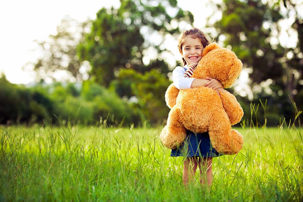 Smiling child standing in a grassy field hugging a large fluffy teddy bear, with trees and sunlight in the background.