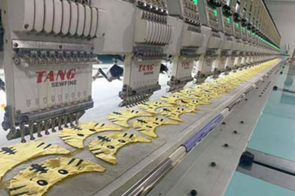Industrial embroidery machines stitching yellow plush fabric pieces with cartoon faces in a plush toy factory production line.