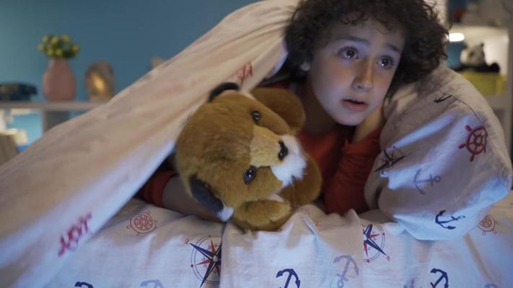 A young boy hiding under a blanket decorated with nautical patterns, holding a brown and white plush dog while looking ahead with curiosity.