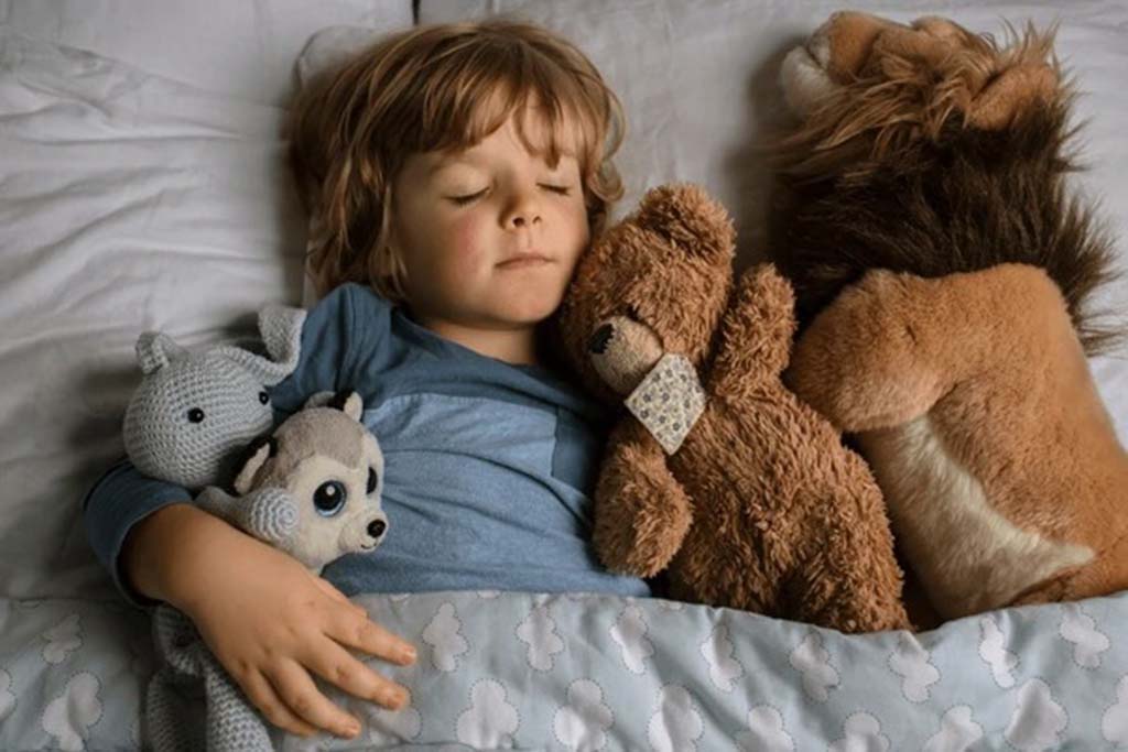  Young child peacefully sleeping in bed, cuddling multiple soft stuffed animals including a teddy bear, lion, and knitted toy under a cloud-patterned blanket.