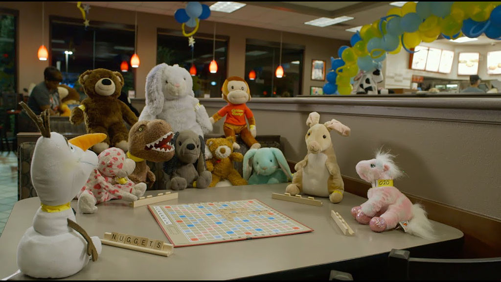Group of plush toys arranged around a table in a restaurant, appearing to play Scrabble under festive balloons and warm lighting