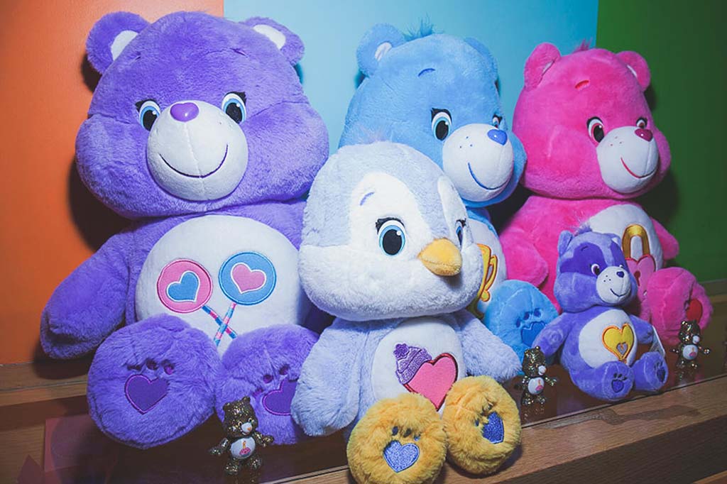Group of large and small plush bears in purple, blue, pink, and lavender colors with heart and balloon designs on their bellies, displayed on a shelf against a colorful background.