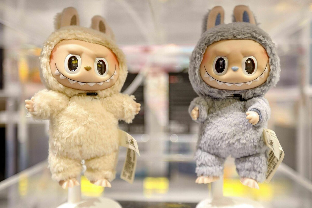 Two collectible plush figures with large eyes and bunny-eared hoodies—one cream-colored and one gray—displayed on white stands in a bright store setting.