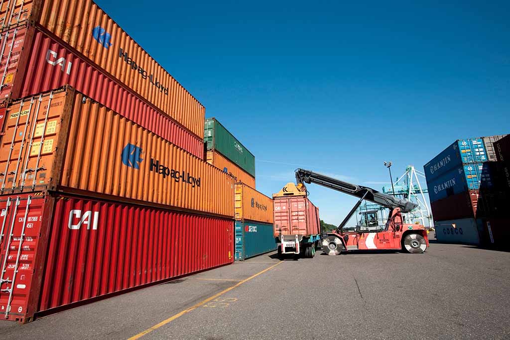 Cargo containers stacked at a shipping port with a crane loading a red container onto a truck under a clear blue sky.