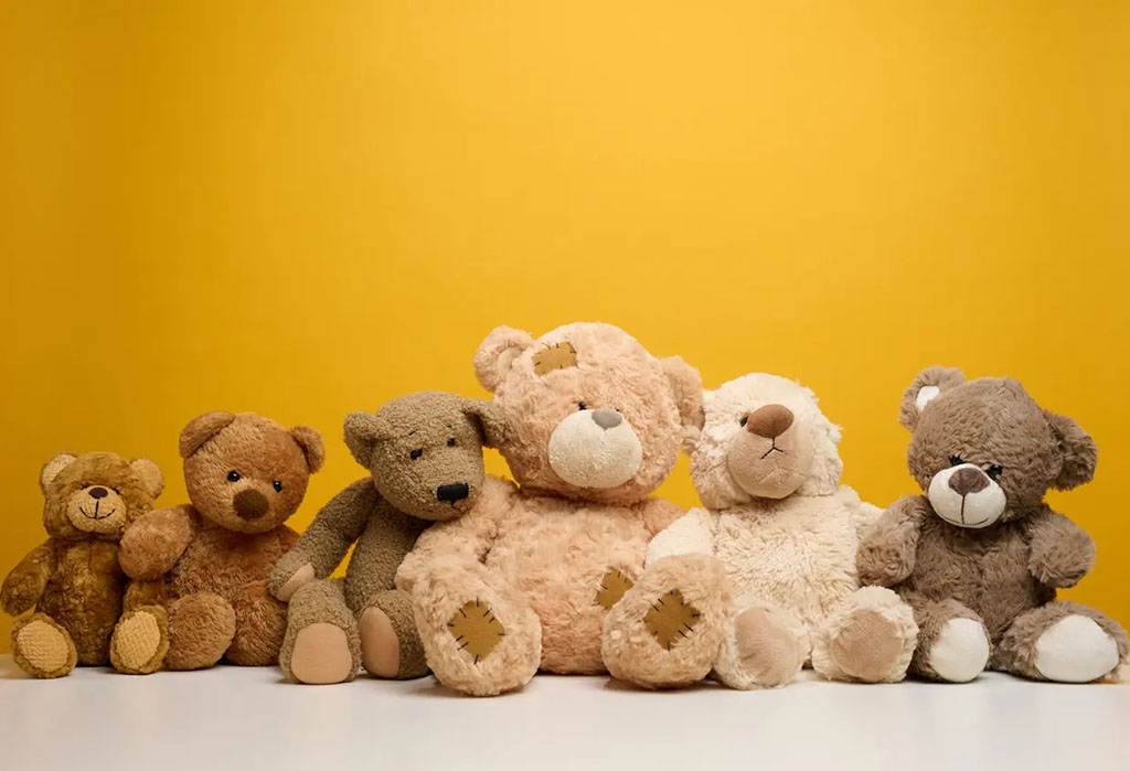 Six teddy bears in different shades of brown and beige sitting in a row against a bright yellow background on a white surface.