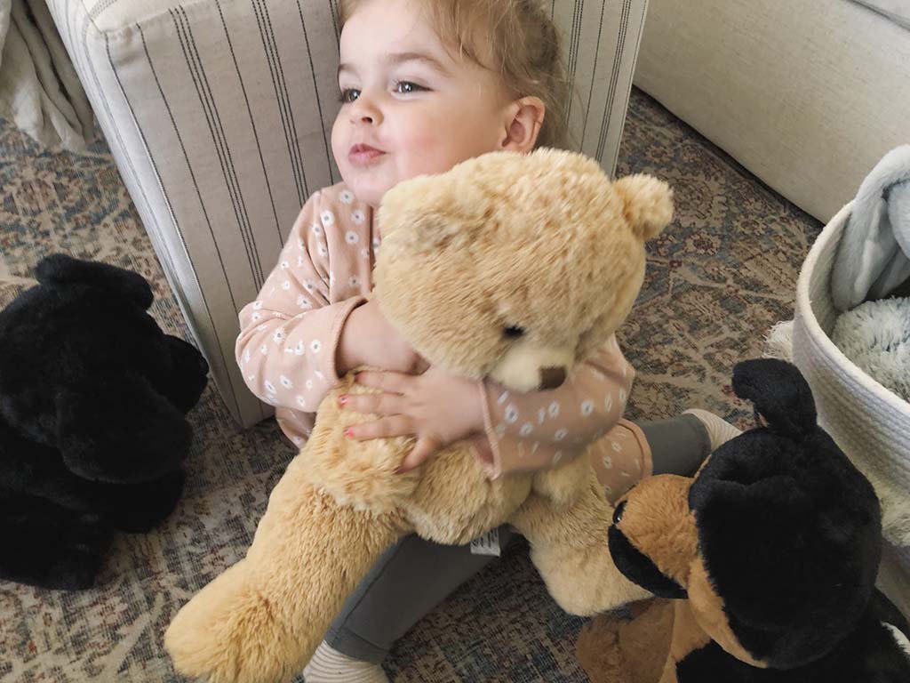 Young child sitting on the floor hugging a light brown teddy bear, surrounded by plush dog toys in a cozy living room.
