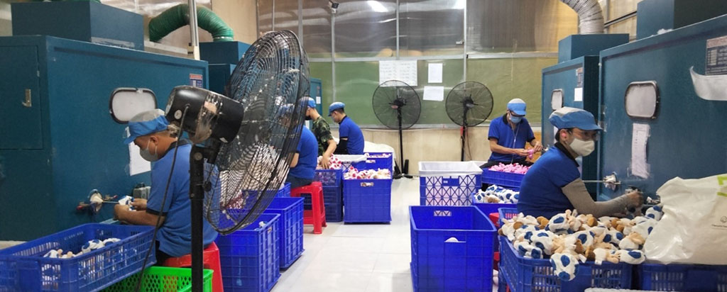 Workers in blue uniforms and masks inspecting stuffed toys in a factory production area.